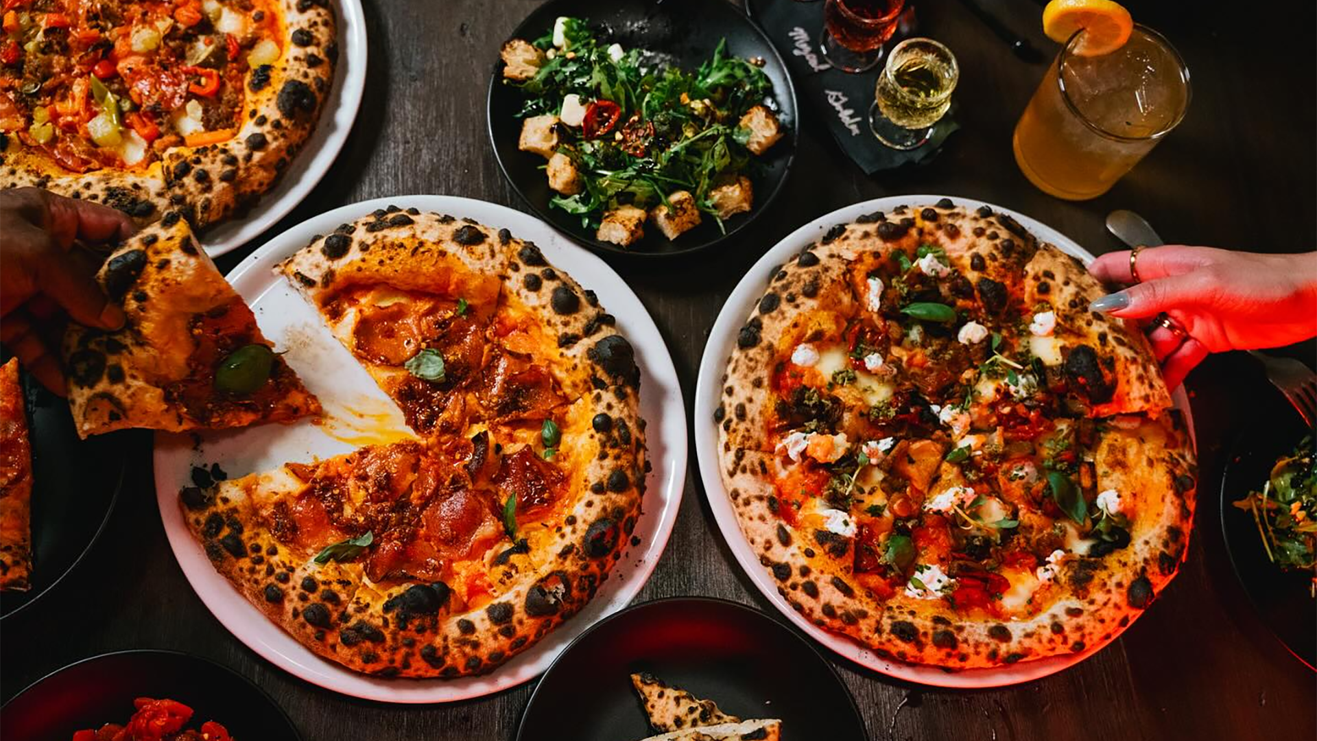 two pizzas with people reaching for slices and salad on the side